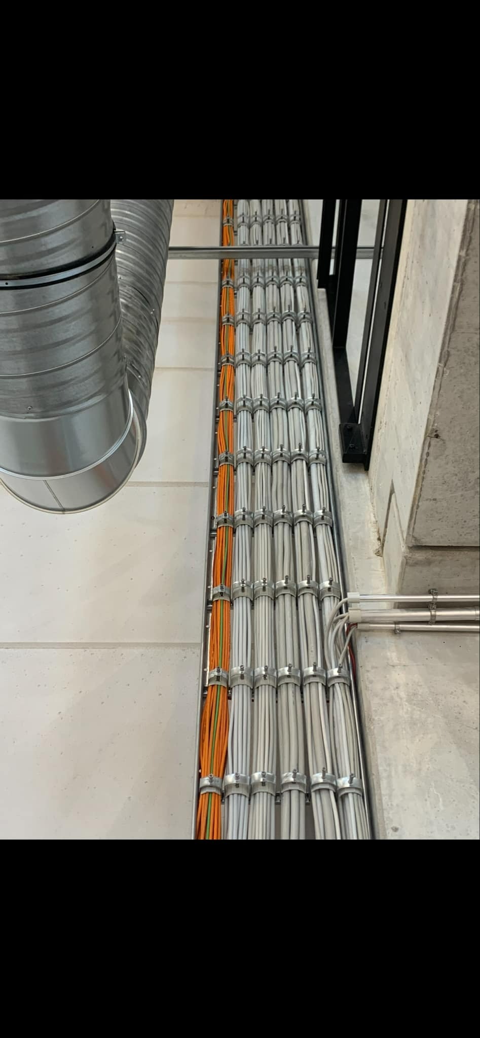 Równolegle ułożone kable elektryczne w korycie, część pomarańczowa, reszta biała, przymocowane metalowymi uchwytami do ściany, widoczny fragment rury wentylacyjnej