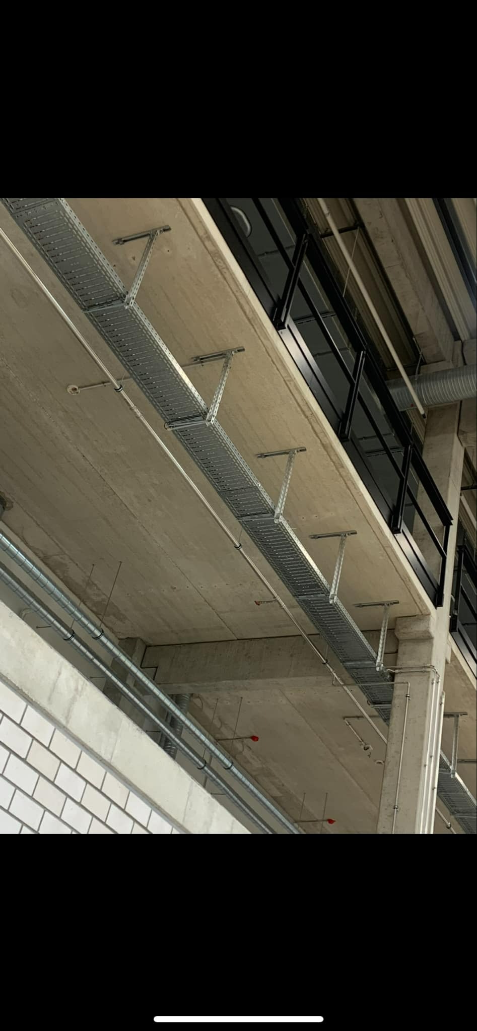 Metalowa korytka kablowa zawieszona pod betonowym sufitem w hali przemysłowej, widoczne rury i instalacje.