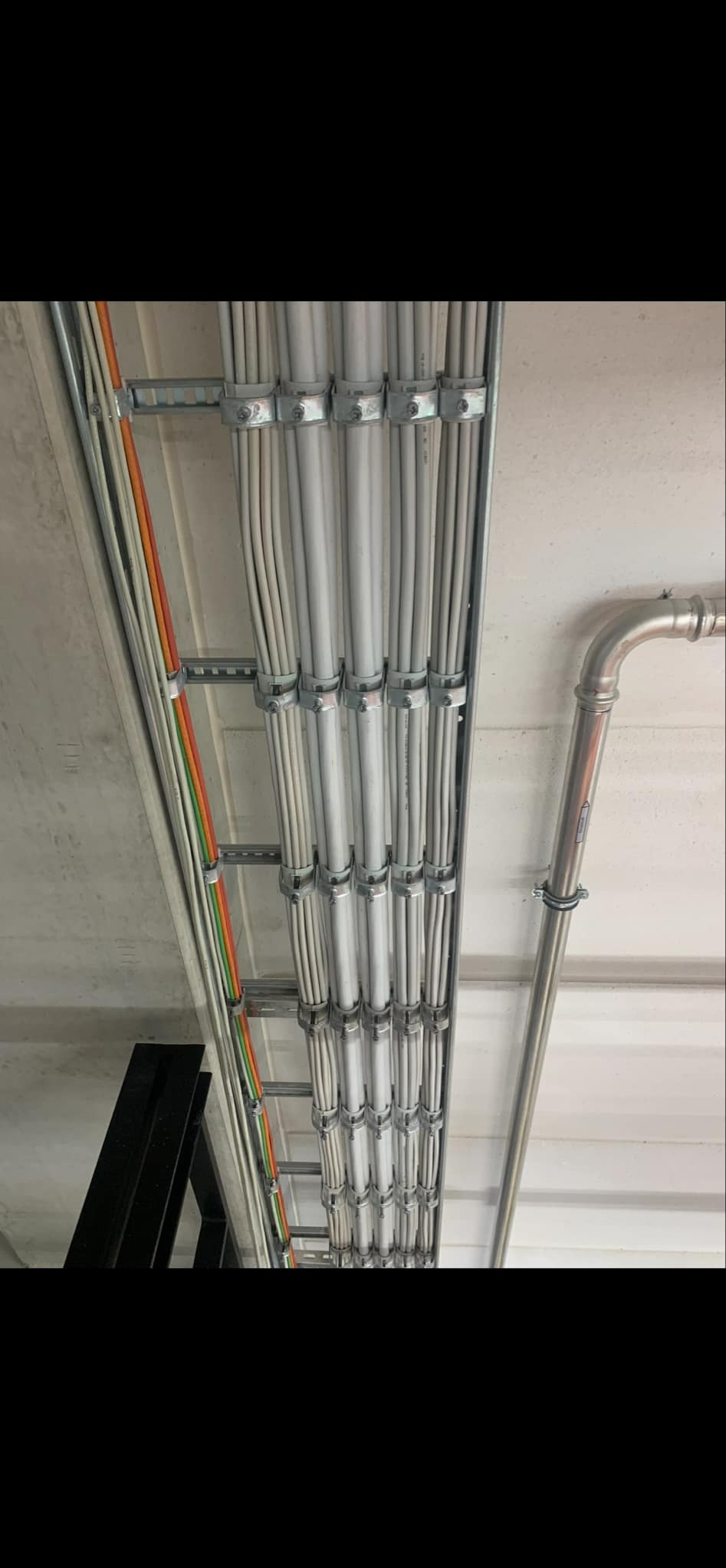 Równoległe wiązki kabli elektrycznych, częściowo kolorowych, mocowane metalowymi uchwytami do drabinki kablowej pod sufitem, obok rura instalacyjna.