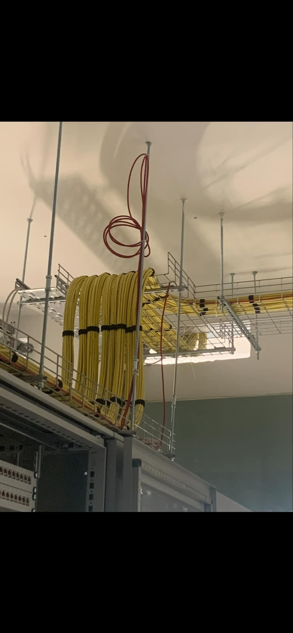 Żółte kable elektryczne ułożone w metalowej korycie pod sufitem, zabezpieczone czarnymi opaskami, z widocznym fragmentem szafy rack