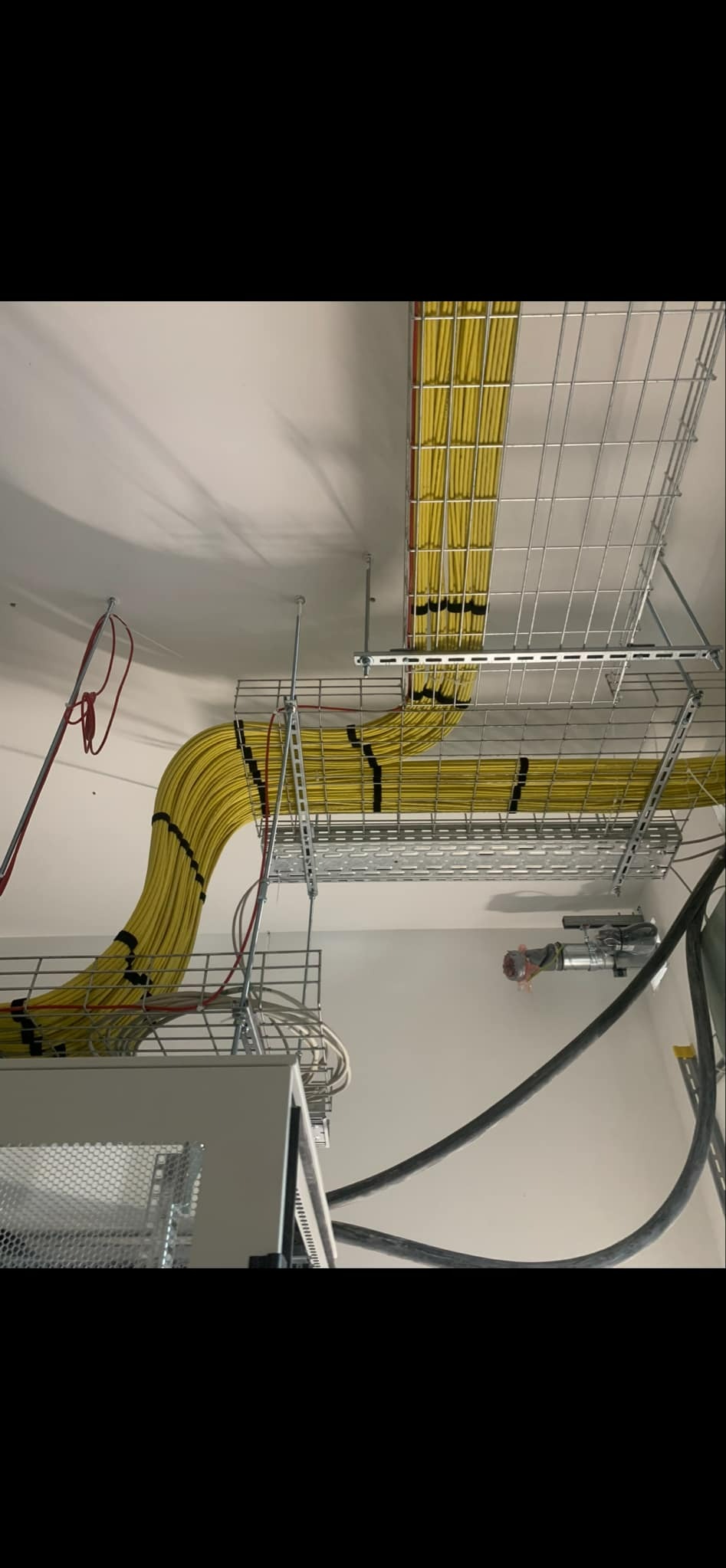 Żółte kable elektryczne ułożone w metalowych korytach pod sufitem, zabezpieczone czarną taśmą, widoczne elementy szafy sterowniczej i inne przewody.