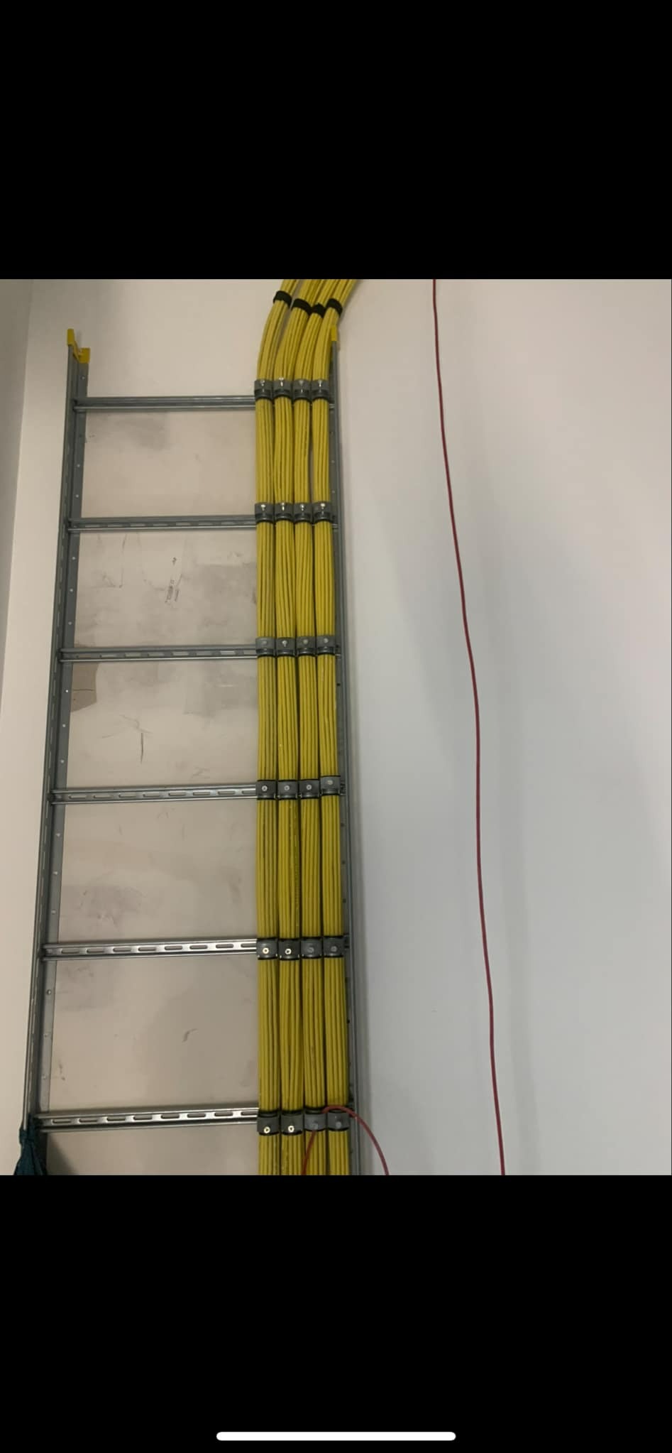 Pionowy, metalowy korytarz kablowy przymocowany do białej ściany, wypełniony gęstą wiązką żółtych przewodów elektrycznych, zabezpieczonych metalowymi uchwytami i czarnymi opaskami zaciskowymi.