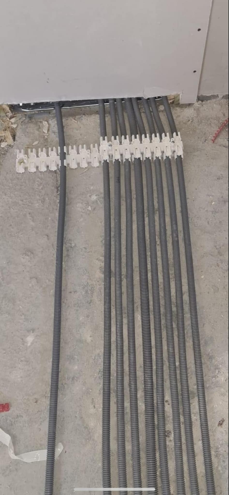 Rury karbowane w szarym kolorze, ułożone równolegle i przymocowane do ściany za pomocą białych uchwytów, widoczne na tle surowej podłogi i ściany w trakcie wykończenia.