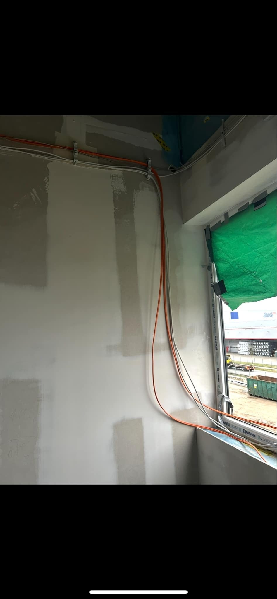 Świeżo położona gładź na ścianie z poprowadzonymi przewodami elektrycznymi, zabezpieczone okno folią ochronną.