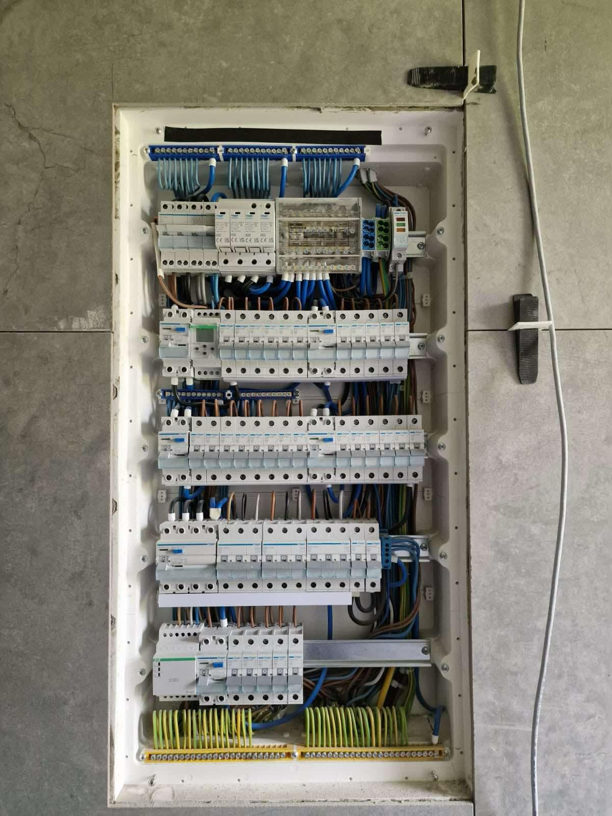 Rozdzielnica elektryczna z widocznymi bezpiecznikami, przewodami w kolorach niebieskim, brązowym i żółto-zielonym, zamontowana na szarej ścianie z płytek ceramicznych, z widocznymi uchwytami...