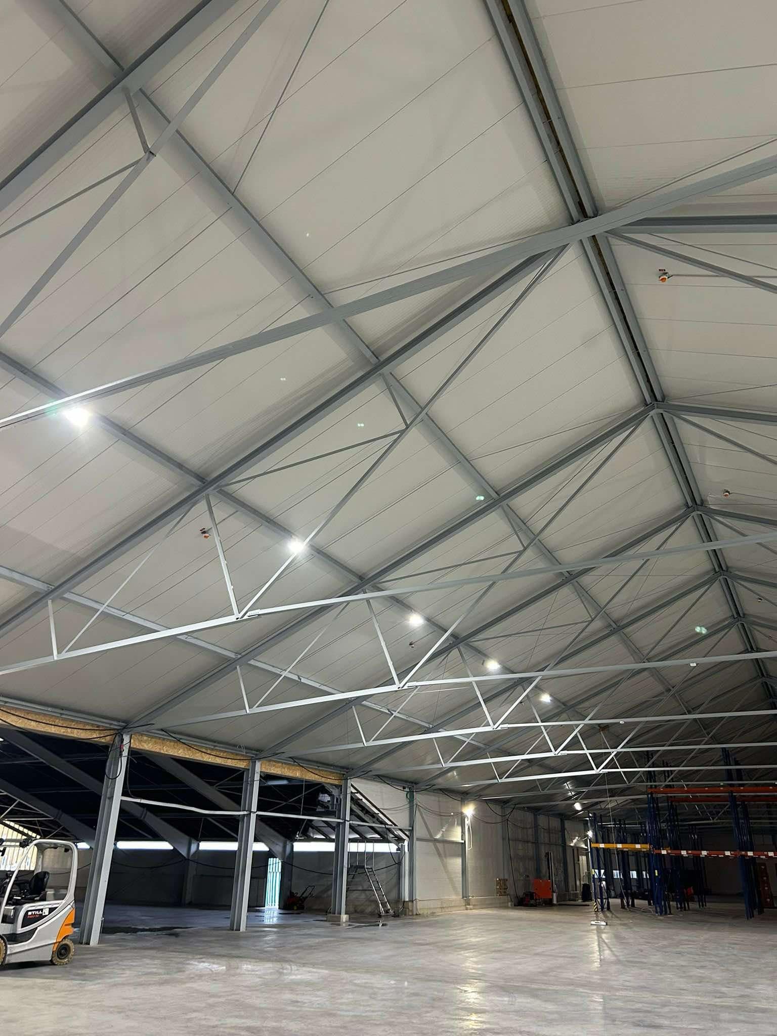 Wnętrze hali magazynowej z widoczną konstrukcją dachu, oświetleniem LED i instalacją przeciwpożarową, w tle wózek widłowy Still.