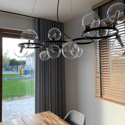 Studio dekoracji okien - Jadalnia z drewnianym stołem, szarymi krzesłami i nowoczesną lampą wiszącą z przezroczystymi kloszami, w tle zasłony i żaluzje drewniane.