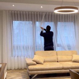 Studio dekoracji okien - Mężczyzna w ciemnej bluzie zawiesza długie, białe, półprzezroczyste zasłony na karniszu w jasnym salonie z beżową sofą i parkietem ułożonym w jodełkę.