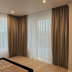 Studio dekoracji okien - Jasny pokój z dużymi oknami przesłoniętymi brązowymi, grubymi zasłonami i białymi, zwiewnymi firanami; widać fragment ramy łóżka.