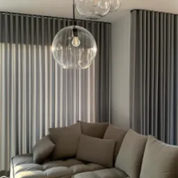 Nowoczesny salon z szarymi, pionowymi zasłonami zaciemniającymi, częściowo przysłoniętymi przez trzy szklane lampy wiszące z żarówkami Edisona, oraz narożną sofą z poduszkami w odcieniach brązu.