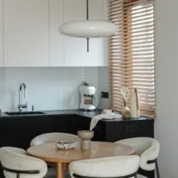 Jasne drewniane żaluzje poziome w oknie nad blatem kuchennym, okrągły stół z krzesłami w minimalistycznej kuchni z białymi szafkami i czarnym blatem.