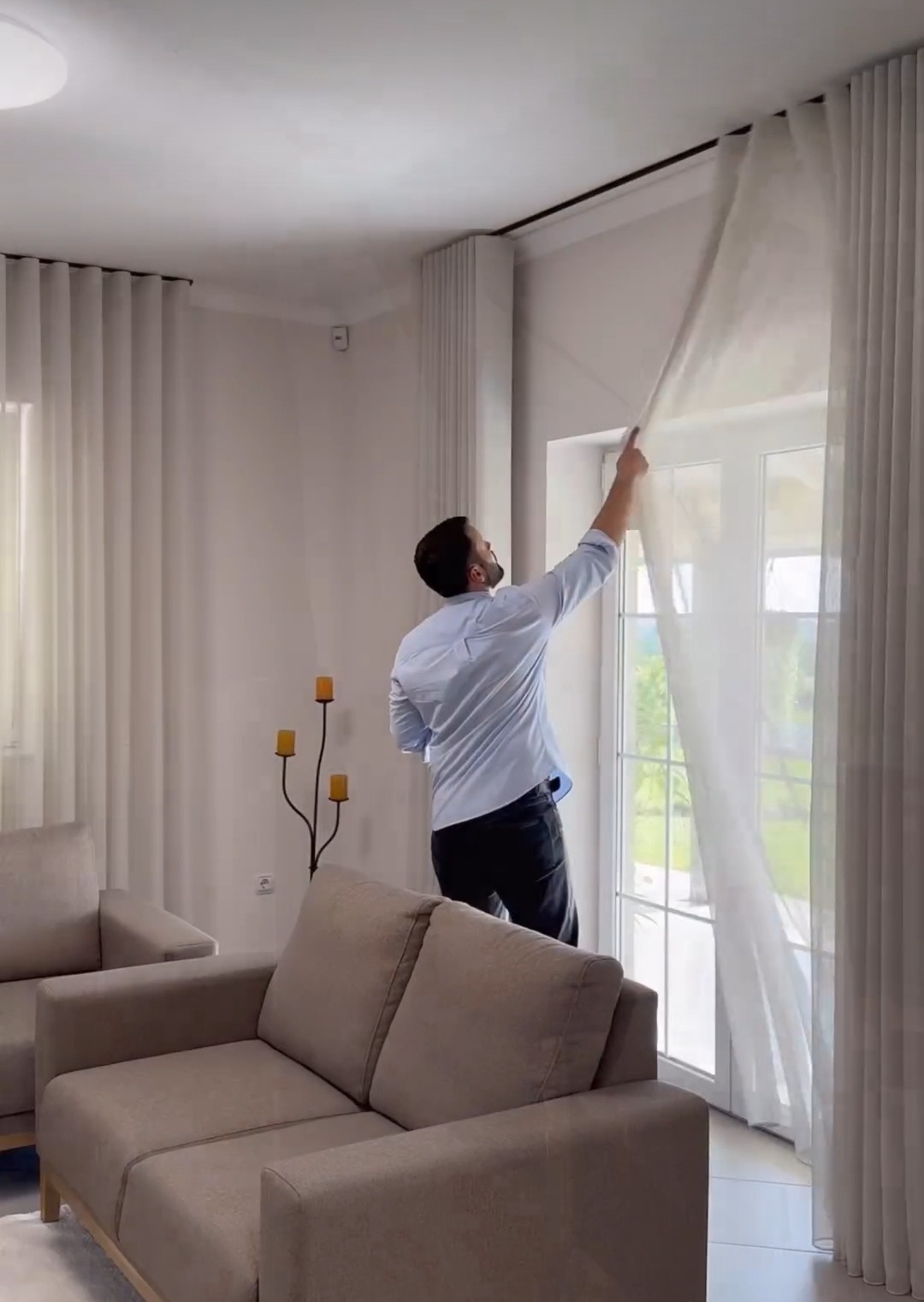 Mężczyzna w jasnej koszuli układa długie, białe, półprzezroczyste zasłony przy oknie w jasnym salonie z beżowymi sofami i ozdobnym świecznikiem.