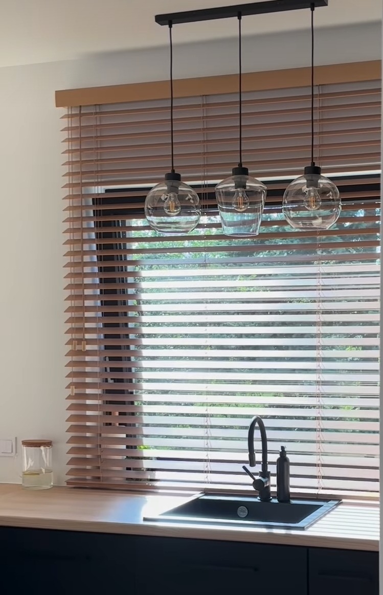 Kuchnia z drewnianymi żaluzjami poziomymi w odcieniach brązu, przez które wpada światło słoneczne, oraz trzema szklanymi lampami wiszącymi nad zlewem.