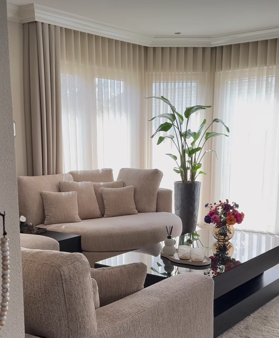 Elegancki salon z beżowymi zasłonami do podłogi, wpuszczającymi naturalne światło, okrągła sofa z poduszkami i duży stolik kawowy z dekoracjami.