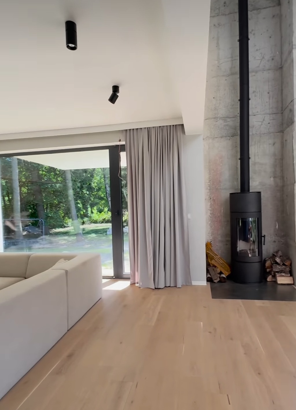 Jasnoszare, gładkie zasłony przy dużym oknie w nowoczesnym salonie z betonową ścianą i kominkiem.