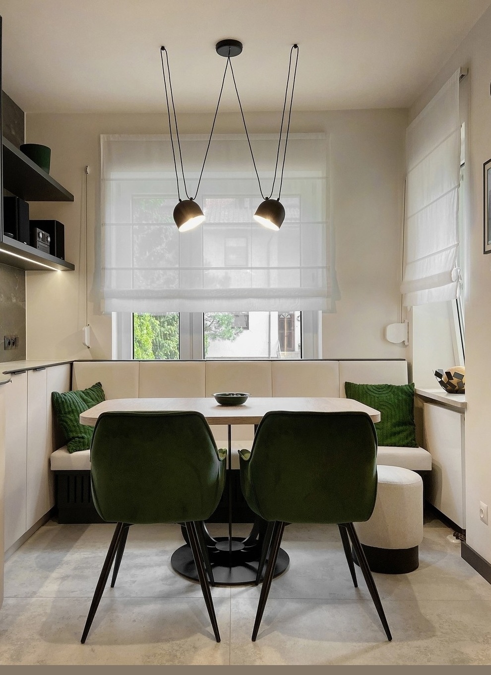 Jasna jadalnia z kremową zabudową, zielonymi poduszkami i krzesłami, oświetlona wiszącymi lampami nad stołem, z oknem przesłoniętym delikatną, białą tkaniną w stylu rolety rzymskiej.