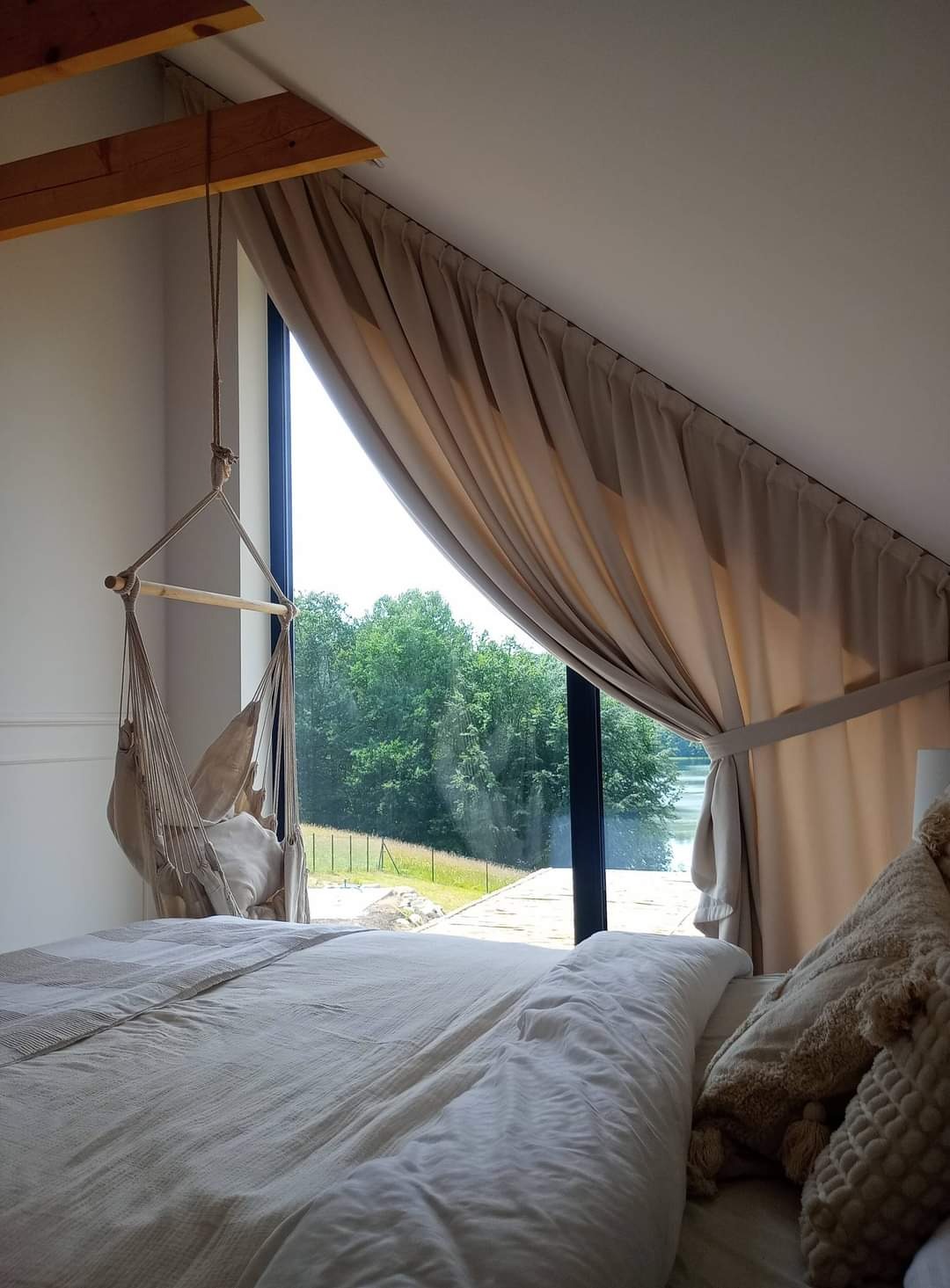 Beżowe, marszczone zasłony przy oknie w sypialni na poddaszu, widok na jezioro i las, hamak wiszący przy oknie.