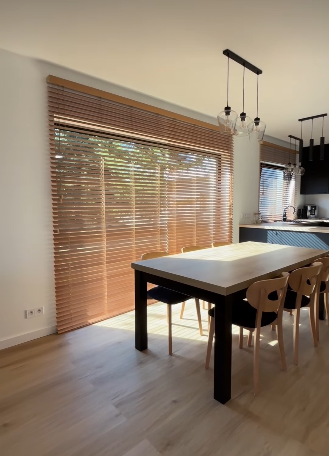 Jasne drewniane żaluzje poziome w salonie z dużym oknem, stół z krzesłami w tle kuchnia z nowoczesnym oświetleniem