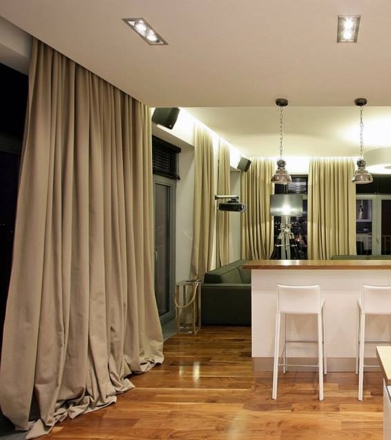 Beżowe, grube zasłony w nowoczesnym salonie z drewnianą podłogą, widoczne lampy sufitowe i krzesła barowe.