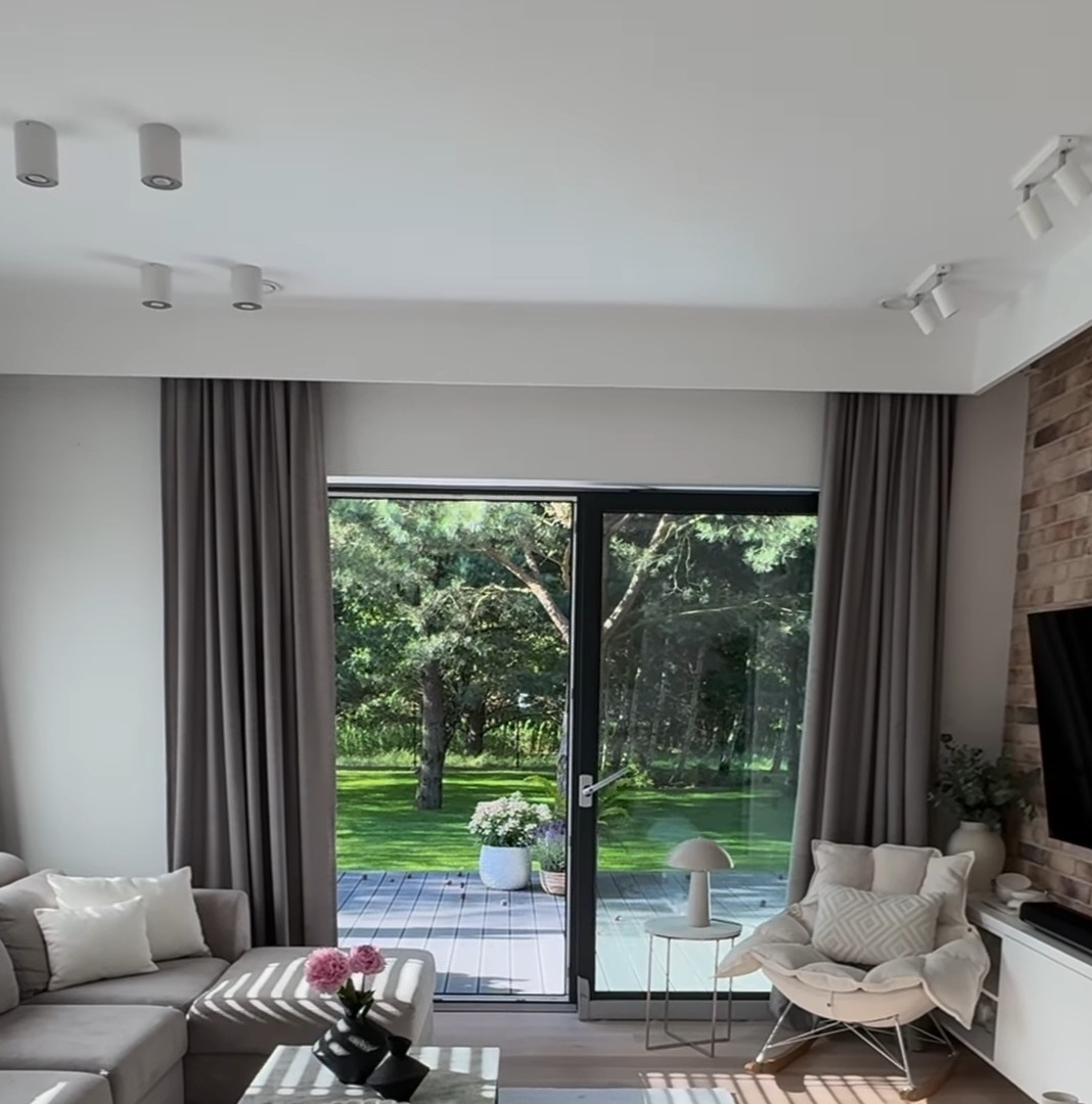 Elegancki salon z szarymi zasłonami po bokach dużego okna tarasowego, widok na zielony ogród z drewnianym tarasem, nowoczesne oświetlenie sufitowe.