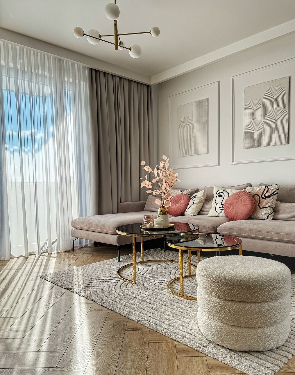 Elegancki salon z beżową sofą, okrągłymi stolikami kawowymi ze złotymi nogami i puszystym dywanem w geometryczne wzory; okno przesłonięte jasną, zwiewną firaną i grubą, beżową tkaniną.