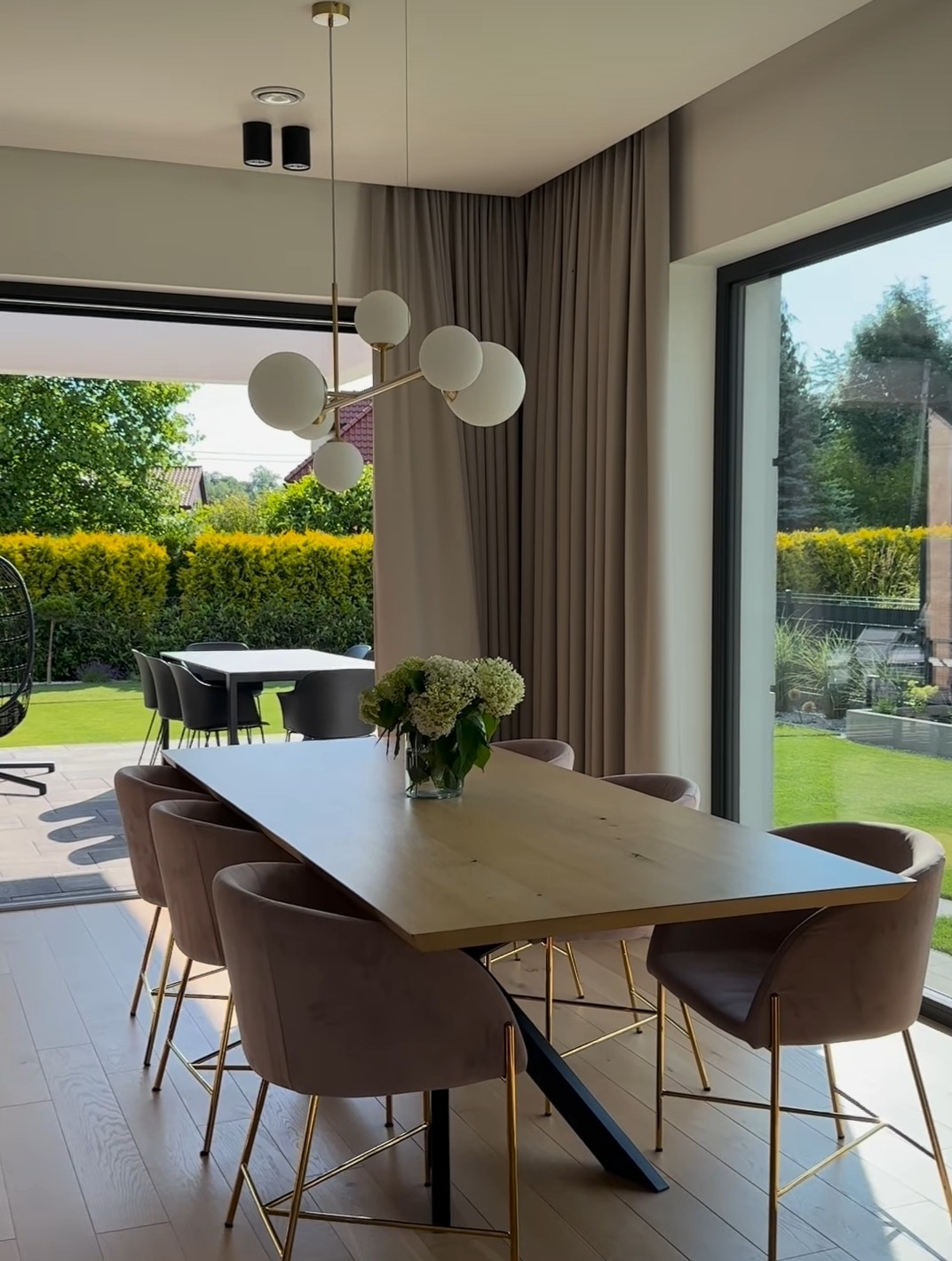 Jasne wnętrze jadalni z dużym oknem na ogród, drewniany stół z bukietem hortensji i krzesłami obitymi beżowym welurem, zasłony w odcieniach szarości.