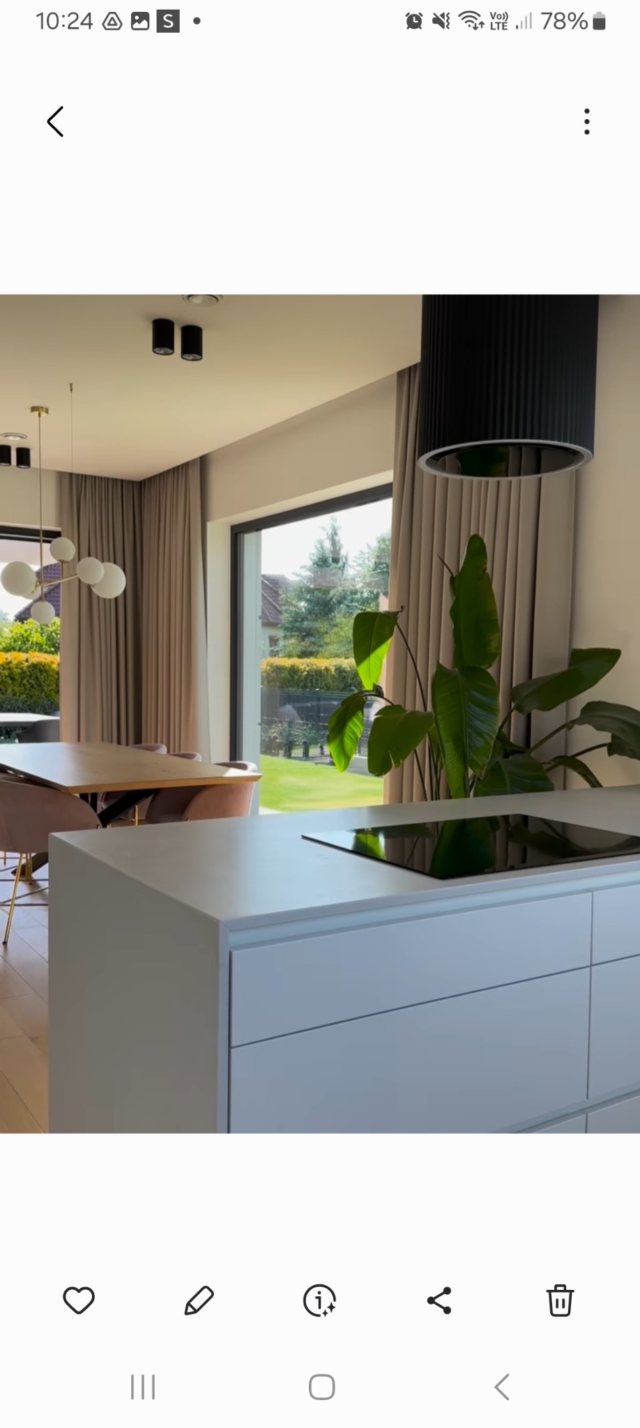 Nowoczesne wnętrze z beżowymi zasłonami, widok na ogród przez duże okno, minimalistyczna lampa i biała wyspa kuchenna.