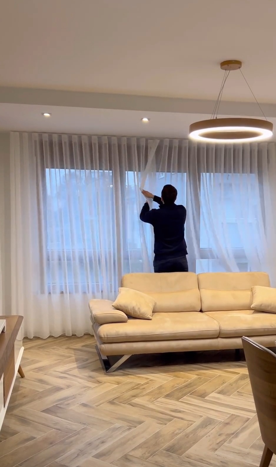 Mężczyzna w ciemnej bluzie zawiesza długie, białe, półprzezroczyste zasłony na karniszu w jasnym salonie z beżową sofą i parkietem ułożonym w jodełkę.