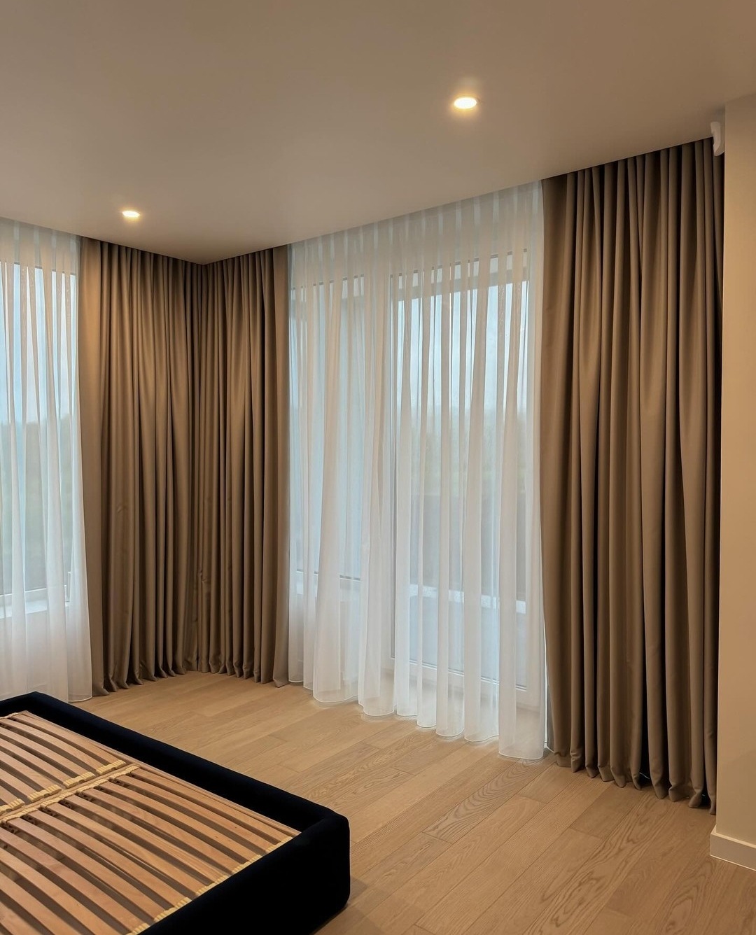 Jasny pokój z dużymi oknami przesłoniętymi brązowymi, grubymi zasłonami i białymi, zwiewnymi firanami; widać fragment ramy łóżka.