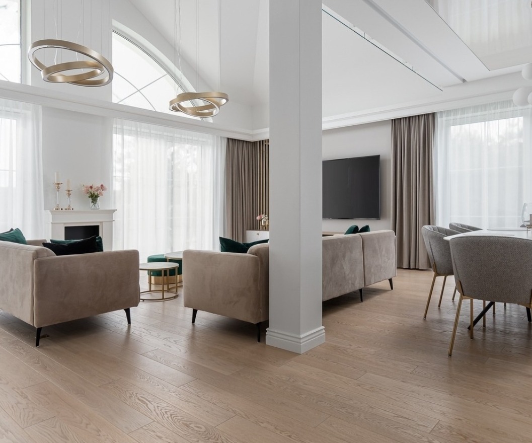 Jasny salon z dużymi oknami przesłoniętymi delikatnymi, białymi zasłonami, beżowe sofy z zielonymi poduszkami, drewniana podłoga i szare krzesła przy stole.