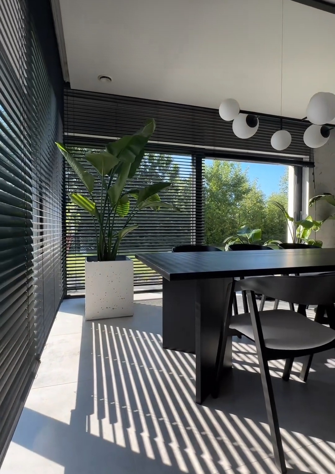 Nowoczesne wnętrze jadalni z czarnym stołem i krzesłami, oświetlone światłem wpadającym przez poziome żaluzje, tworzące geometryczne cienie na podłodze, z rośliną w białej donicy.