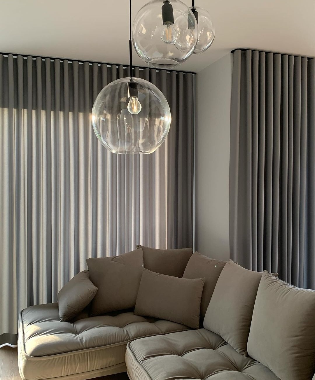 Nowoczesny salon z szarymi, pionowymi zasłonami zaciemniającymi, częściowo przysłoniętymi przez trzy szklane lampy wiszące z żarówkami Edisona, oraz narożną sofą z poduszkami w odcieniach brązu.