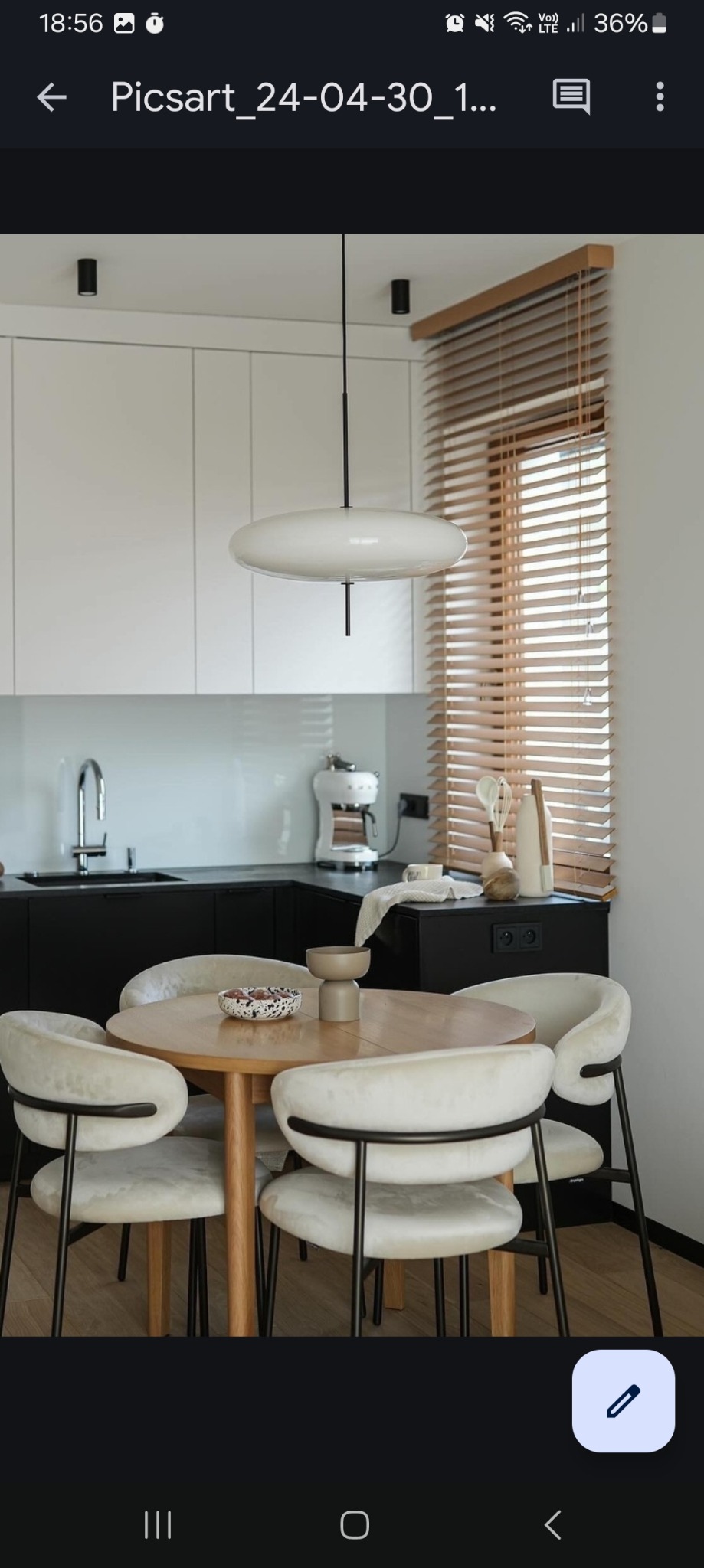 Jasne drewniane żaluzje poziome w oknie nad blatem kuchennym, okrągły stół z krzesłami w minimalistycznej kuchni z białymi szafkami i czarnym blatem.