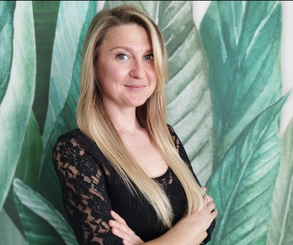 Portret uśmiechniętej kobiety z blond włosami w czarnej koronkowej bluzce, skrzyżowane ręce na tle ściany z motywem liści.