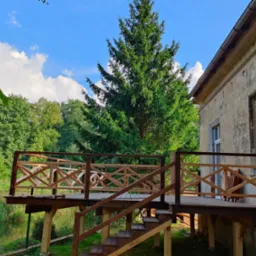 Drewniany taras z balustradą w stylu rustykalnym, przylegający do starego budynku z widoczną cegłą i zielenią. Zbliżenie na konstrukcję tarasu.