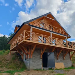 Dwupiętrowy dom z drewna na zboczu, z balkonami z drewnianą balustradą. Widoczne szare bloczki fundamentowe i otwarte drzwi wejściowe, błękitne niebo w tle.