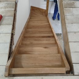 Grzegorz Bielski - Montaż drewnianych schodów wewnętrznych z jasnego drewna, widok z góry na stopniach, w tle okno i fragment osoby w niebieskim kombinezonie.
