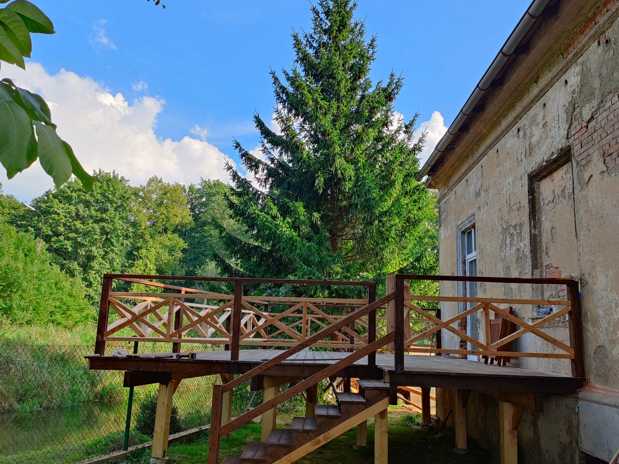 Drewniany taras z balustradą w stylu rustykalnym, przylegający do starego budynku z widoczną cegłą i zielenią. Zbliżenie na konstrukcję tarasu.