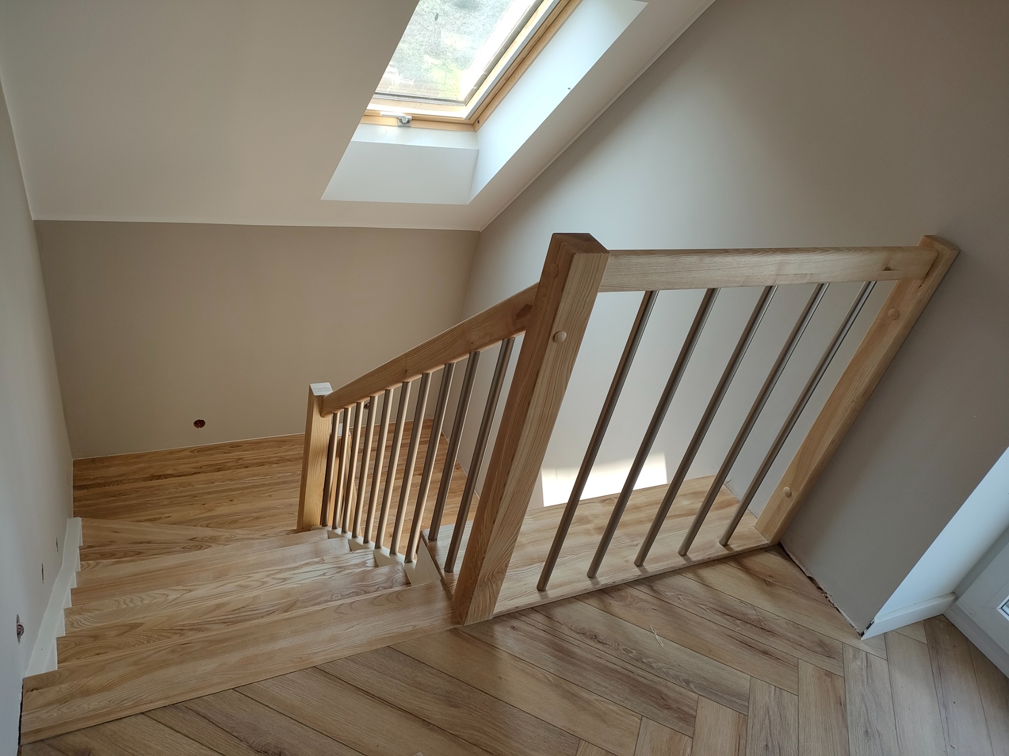 Jasne drewniane schody z metalowymi balustradami w nowoczesnym wnętrzu z oknem dachowym. Parkiet ułożony w jodełkę.