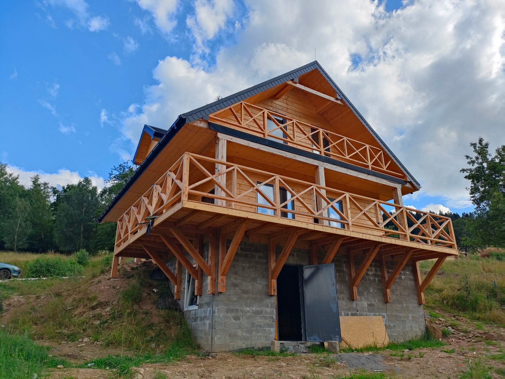 Dwupiętrowy dom z drewna na zboczu, z balkonami z drewnianą balustradą. Widoczne szare bloczki fundamentowe i otwarte drzwi wejściowe, błękitne niebo w tle.