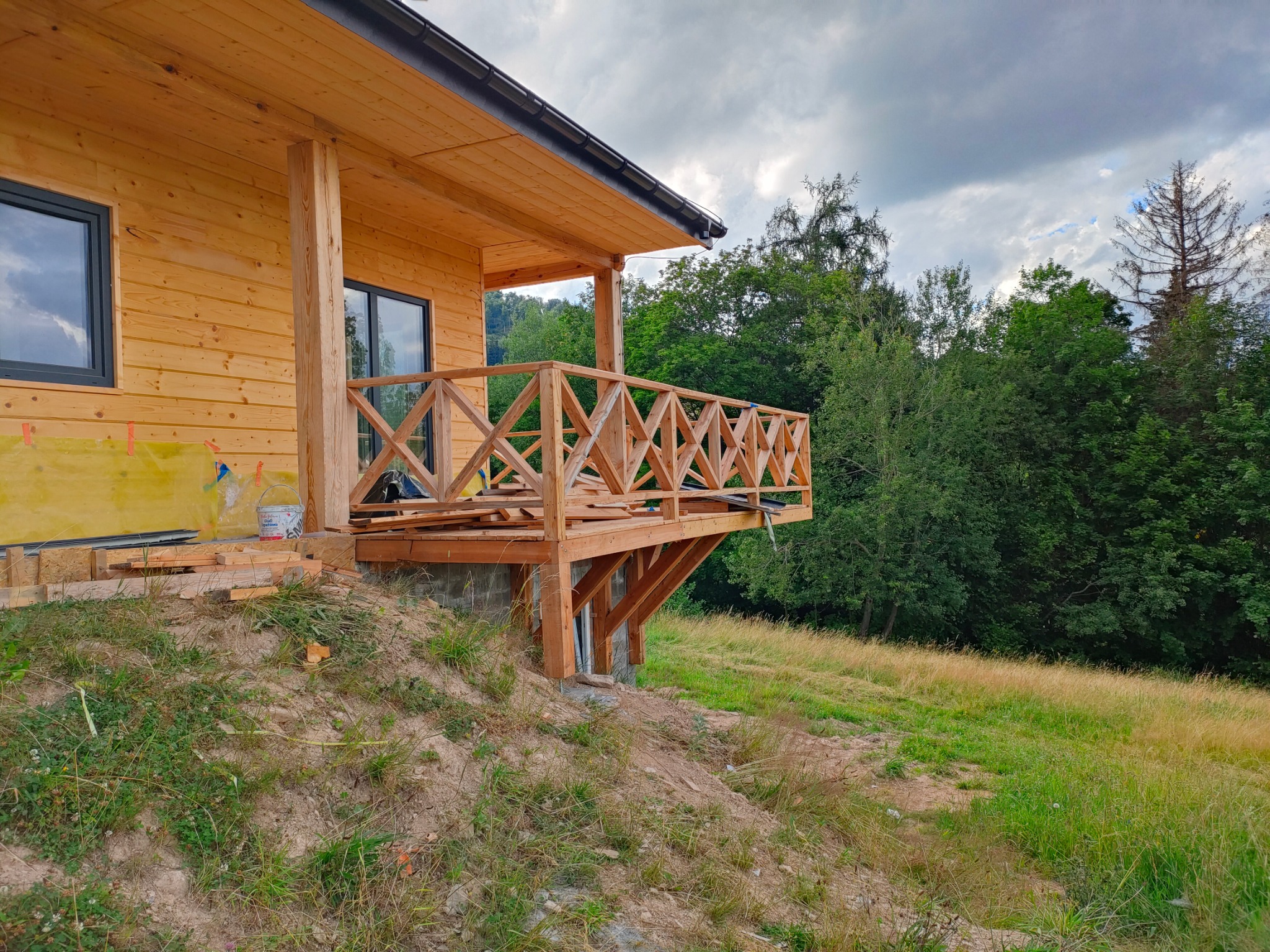 Wznoszący się drewniany taras z balustradą o ażurowym wzorze, częściowo wykończony, przy domku z bali na tle zieleni i pochmurnego nieba.