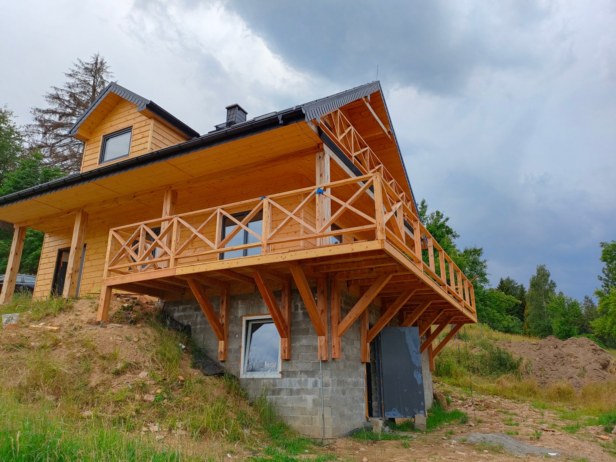 Drewniany dom z tarasem na zboczu, widok z dołu, pochmurne niebo. Konstrukcja z surowego drewna, szara podmurówka, zielone otoczenie.