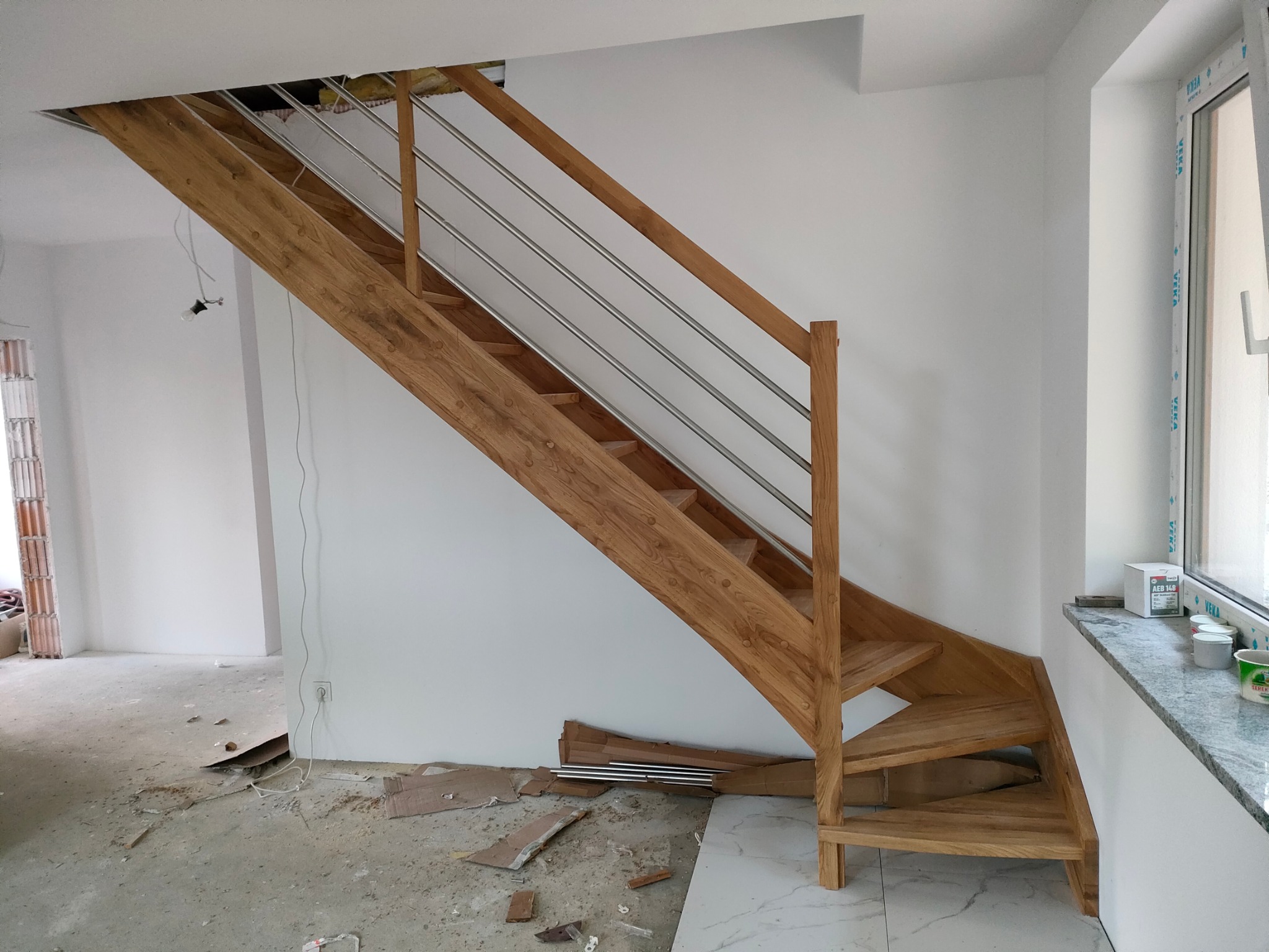 Jasne, drewniane schody z metalową balustradą w surowym wnętrzu. Widoczne niedokończone podłogi i ściany, sugerujące etap wykończeniowy prac budowlanych.