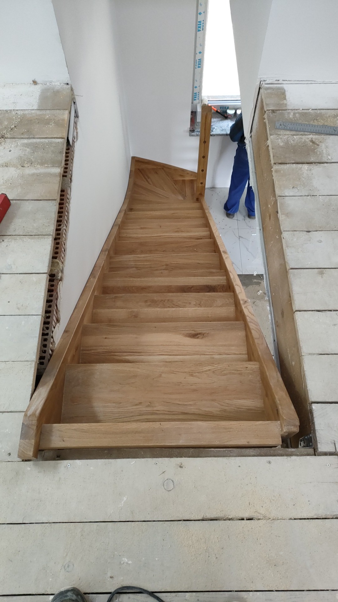 Montaż drewnianych schodów wewnętrznych z jasnego drewna, widok z góry na stopniach, w tle okno i fragment osoby w niebieskim kombinezonie.