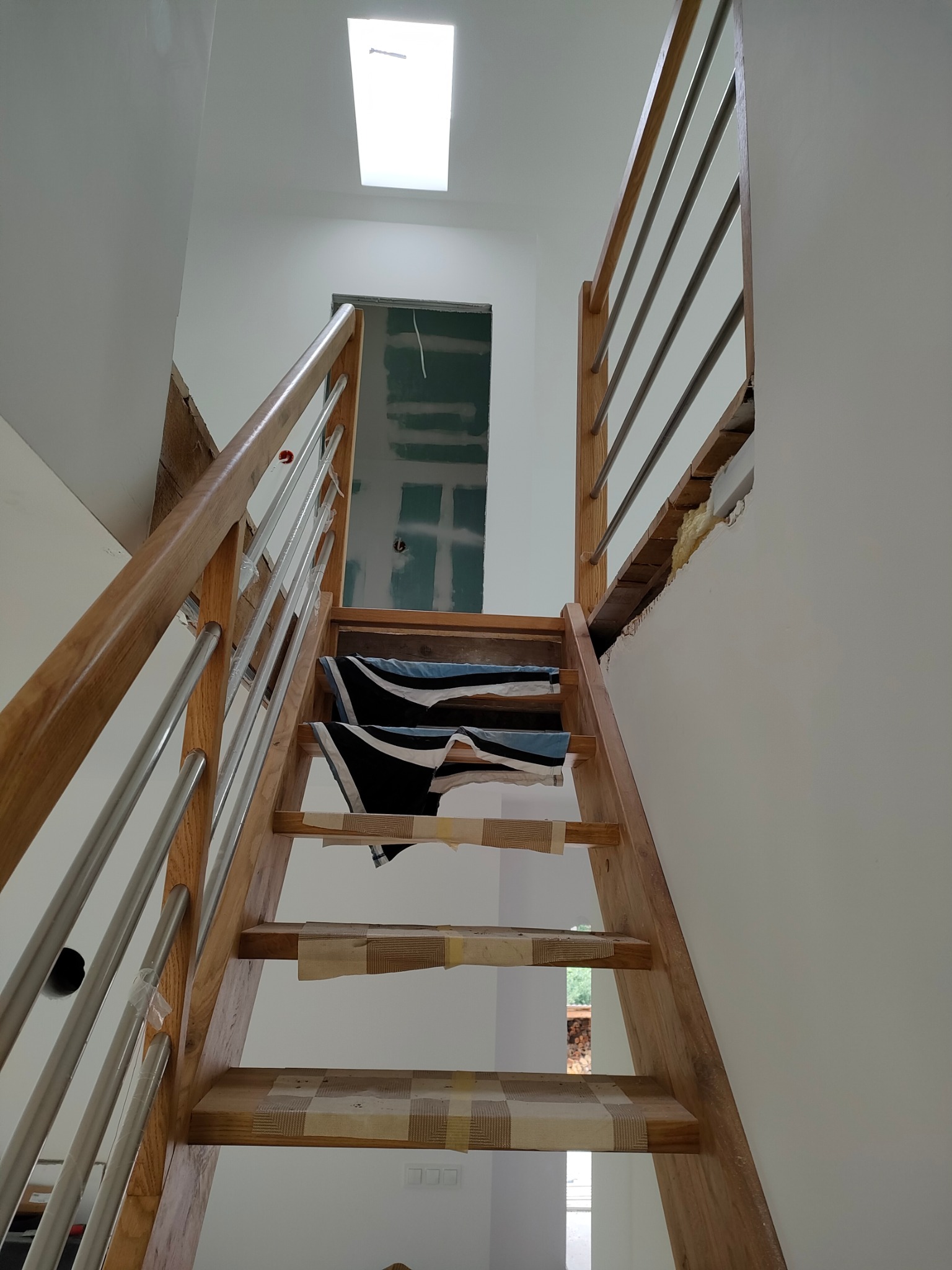 Drewniane schody z metalową balustradą w trakcie montażu, zabezpieczone taśmą. Widoczne niedokończone prace wykończeniowe ścian i sufitu.