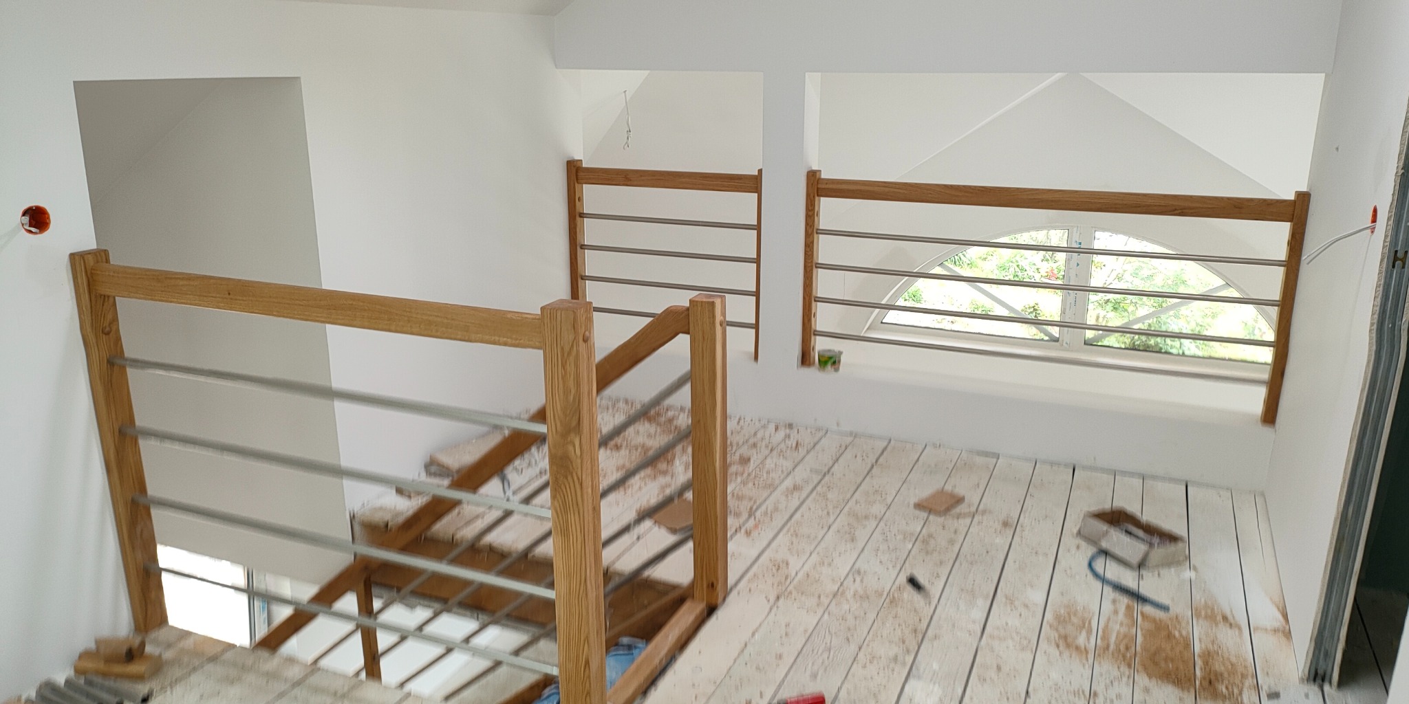 Jasne drewniane schody z metalowymi barierkami w nowoczesnym wnętrzu. Widok z góry na częściowo wykończone schody i podłogę, okno w tle.