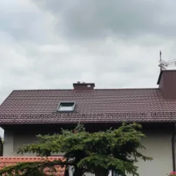 Dom z brązowym dachem z blachodachówki, widoczne okno dachowe, rynny, komin z anteną na tle pochmurnego nieba. Część domu zasłania świerk.