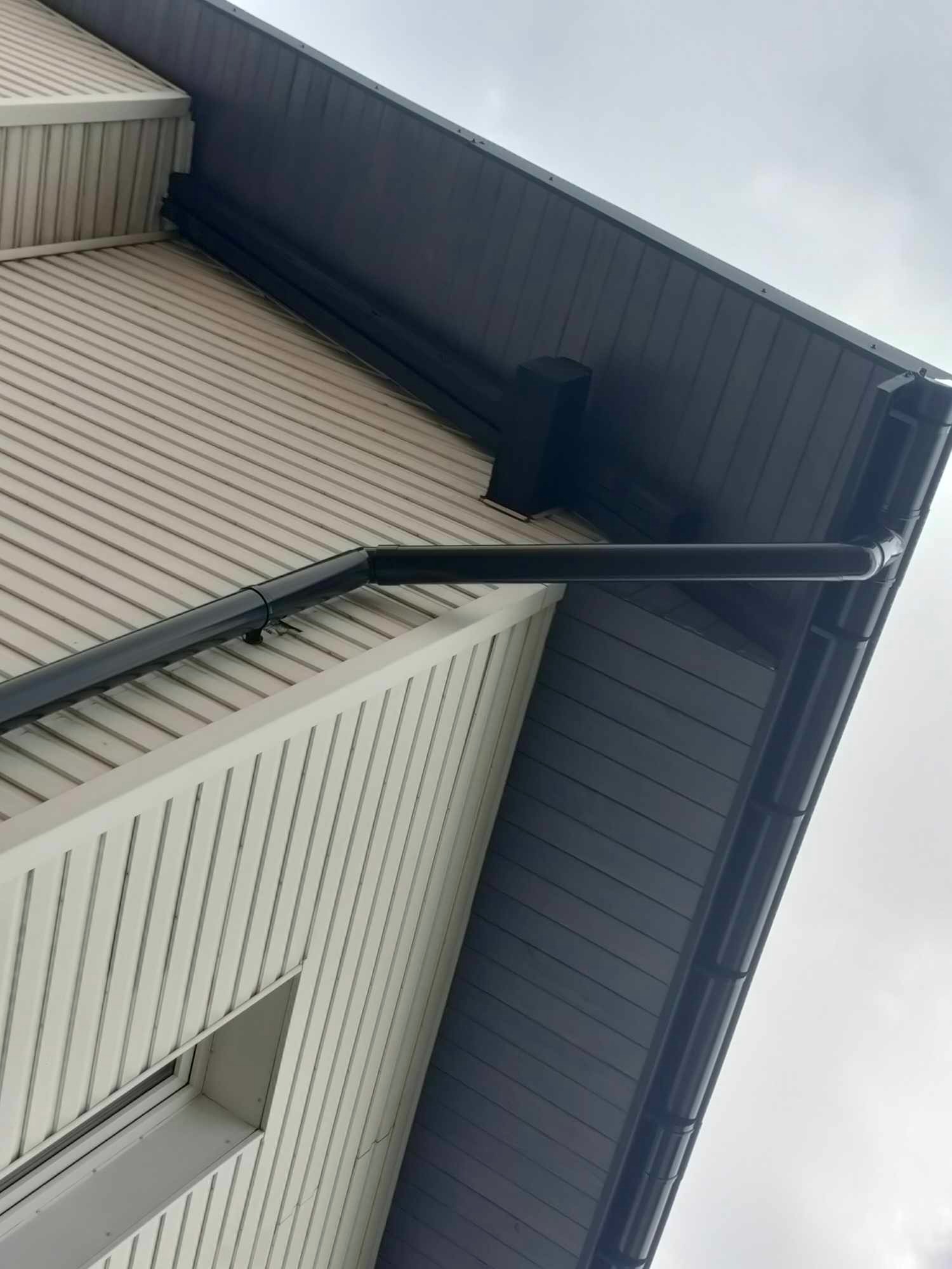 Ciemna rynna spustowa zamontowana na jasnej elewacji domu z widocznym fragmentem dachu krytego ciemną blachodachówką, ujęcie z dołu.