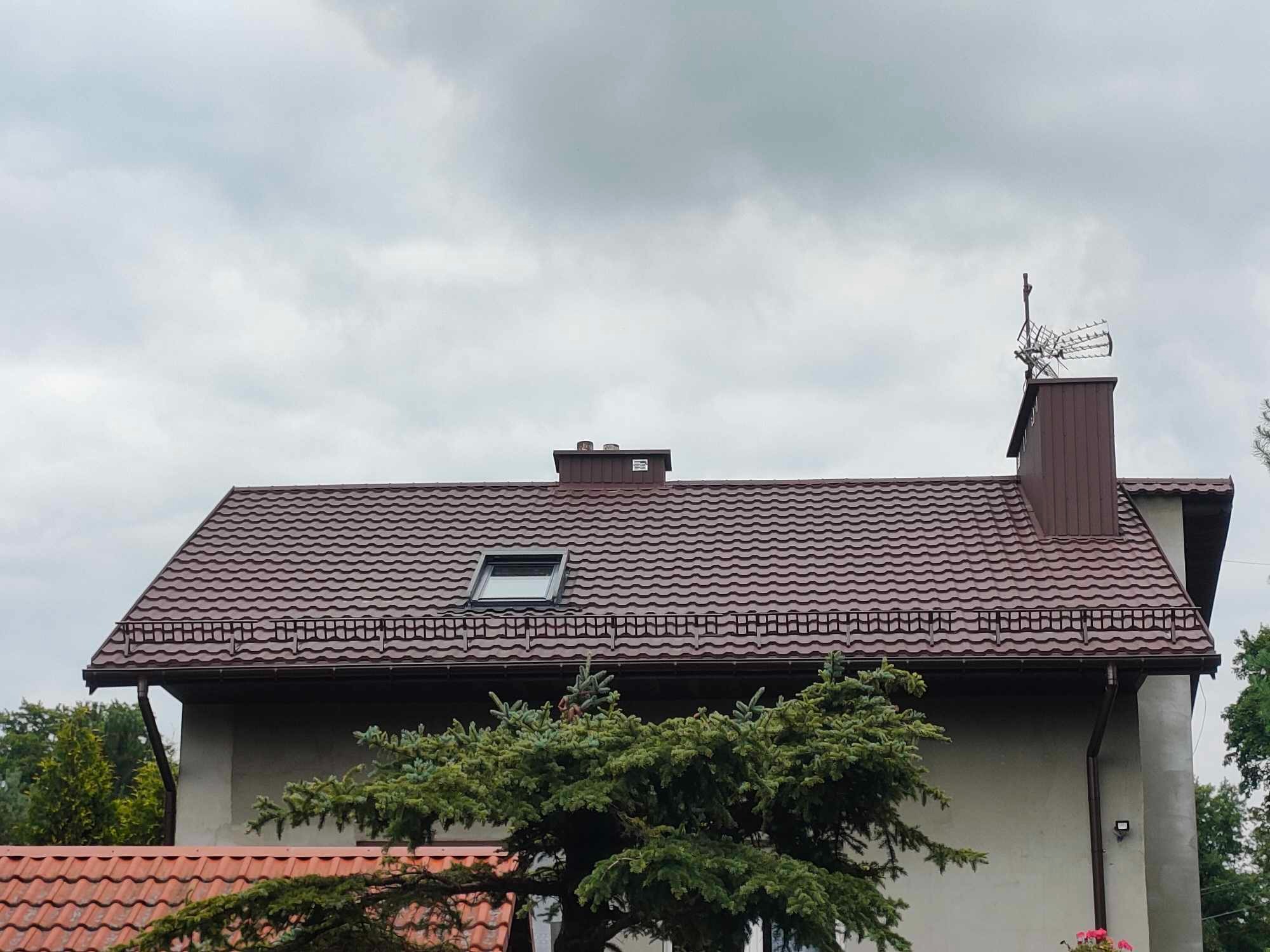 Dom z brązowym dachem z blachodachówki, widoczne okno dachowe, rynny, komin z anteną na tle pochmurnego nieba. Część domu zasłania świerk.