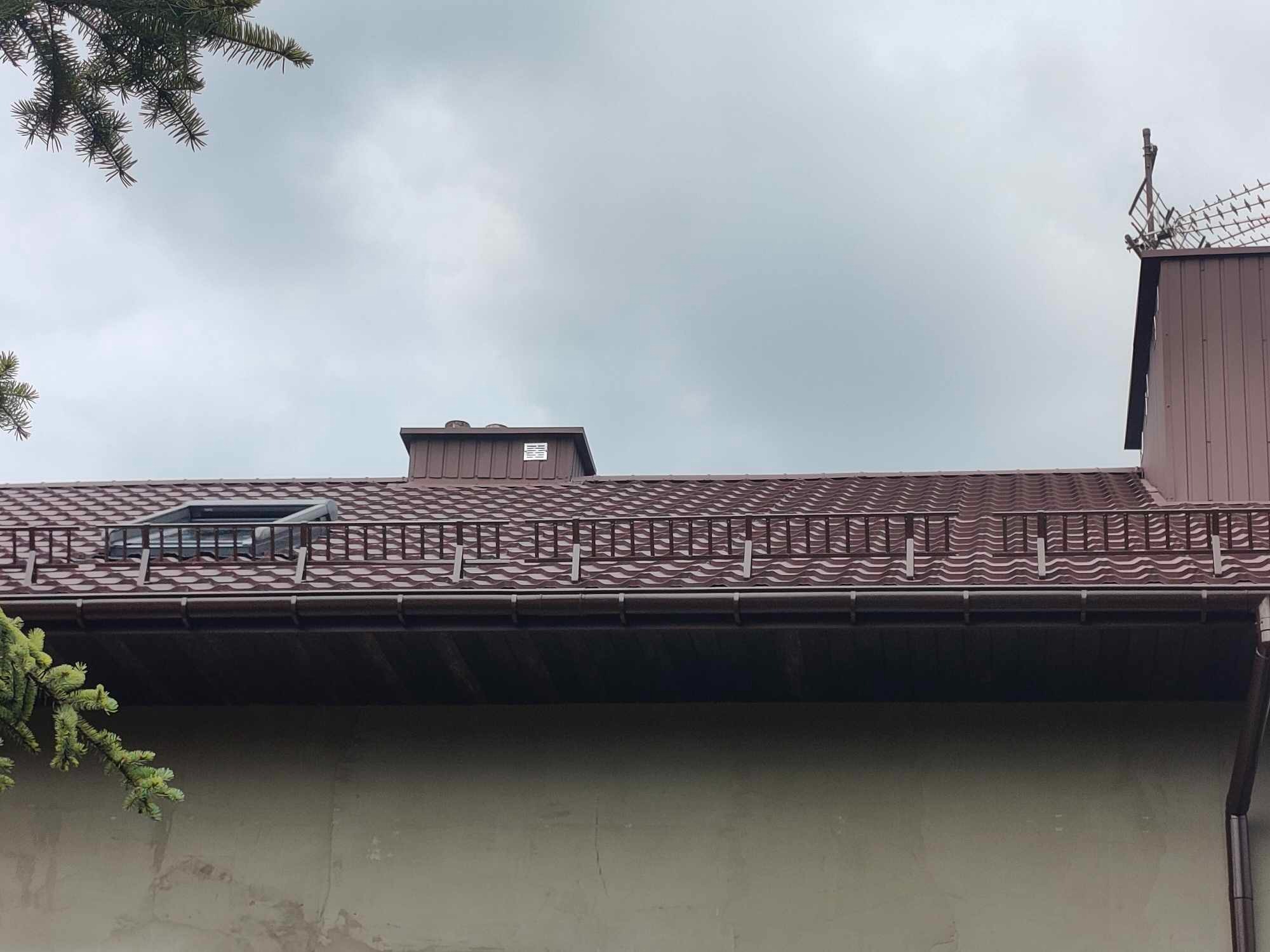 Czerwony dach z blachy falistej z oknem dachowym i ozdobną barierką, widok z dołu na tle pochmurnego nieba.